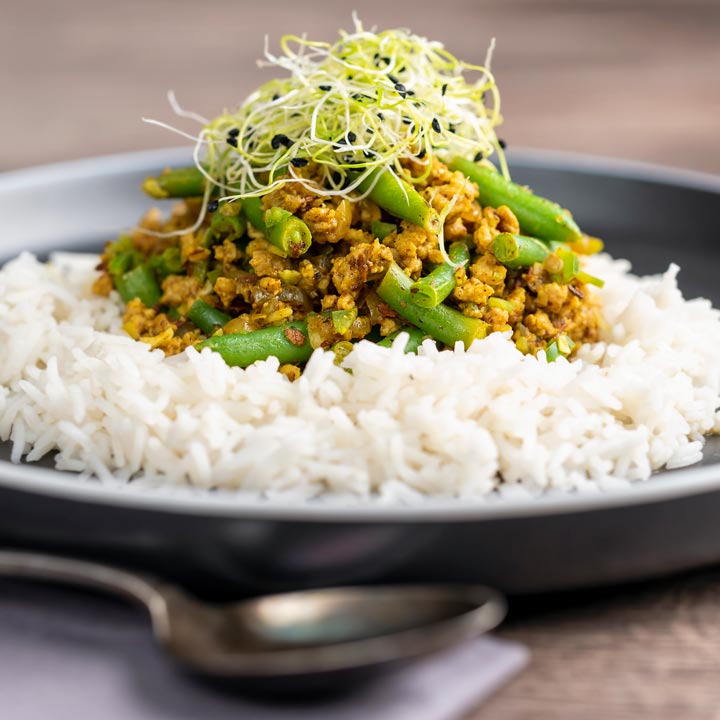 Square image of a pork mince curry with green beans served with boiled rice and sprouted onion seeds on a black plate