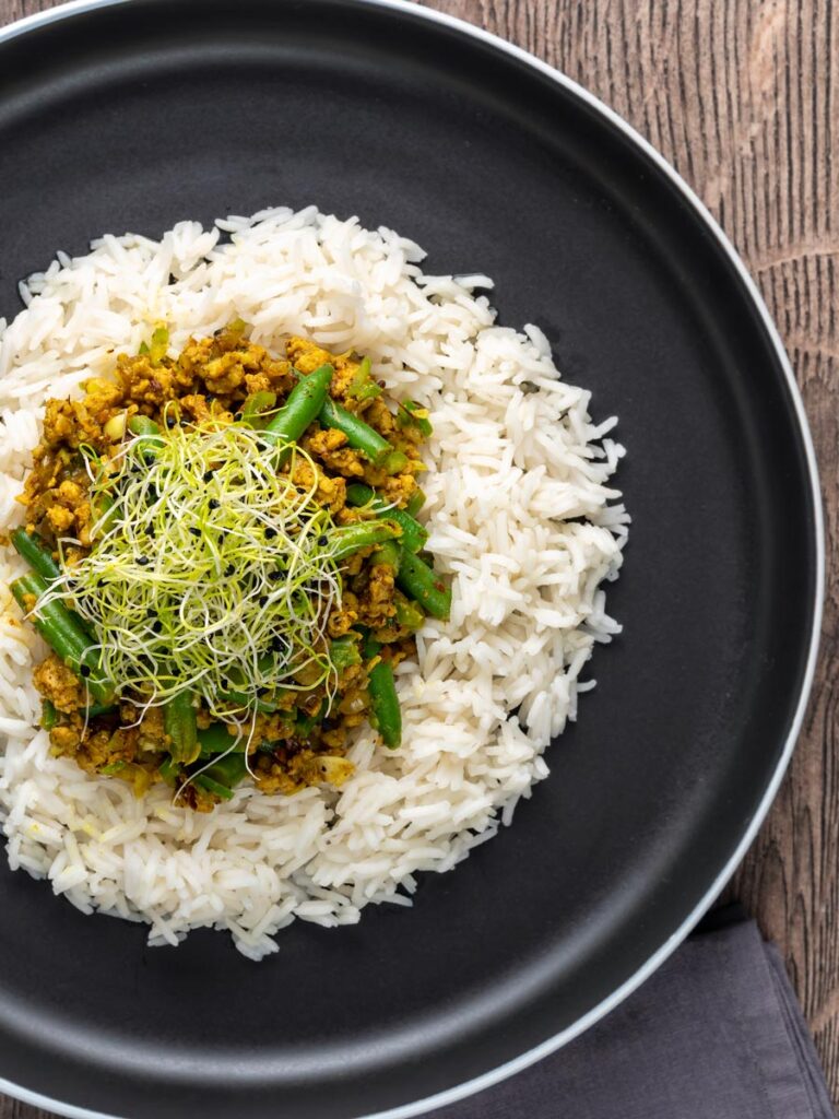 Portrait overhead image of a pork mince curry with green beans served with boiled rice and sprouted onion seeds on a black plate