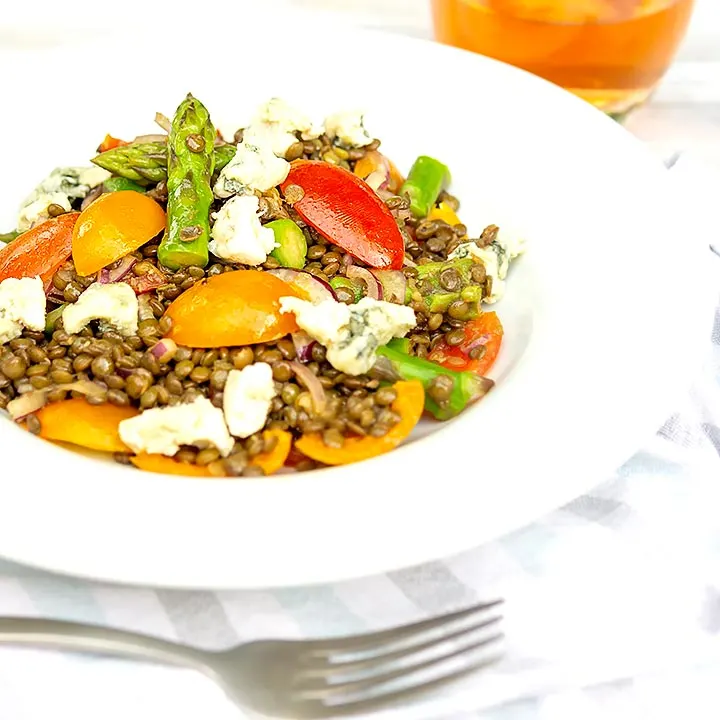 Square image of an asparagus salad with lentils, blue cheese and tomatoes served in a white bowl