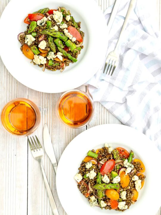 portrait overhead image of an asparagus salad with lentils, blue cheese and tomatoes served in two white bowls served with a glass of wine
