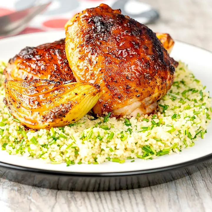 Square image of roasted harissa chicken thighs with onion wedges served on herbed couscous