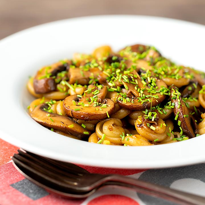 Square image of balsamic mushroom pasta using volanti served with snipped chives in a white bowl