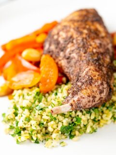 Portrait image of a rabbit leg tagine with almonds and dried apricots served with herby bulgur wheat on a white plate