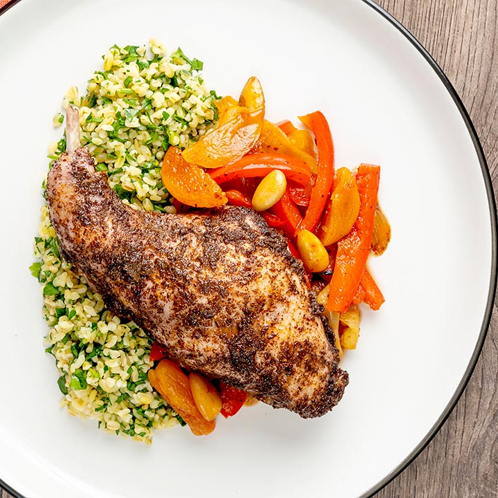 Square overhead image of a rabbit leg tagine with almonds and dried apricots served with herby bulgur wheat on a white plate