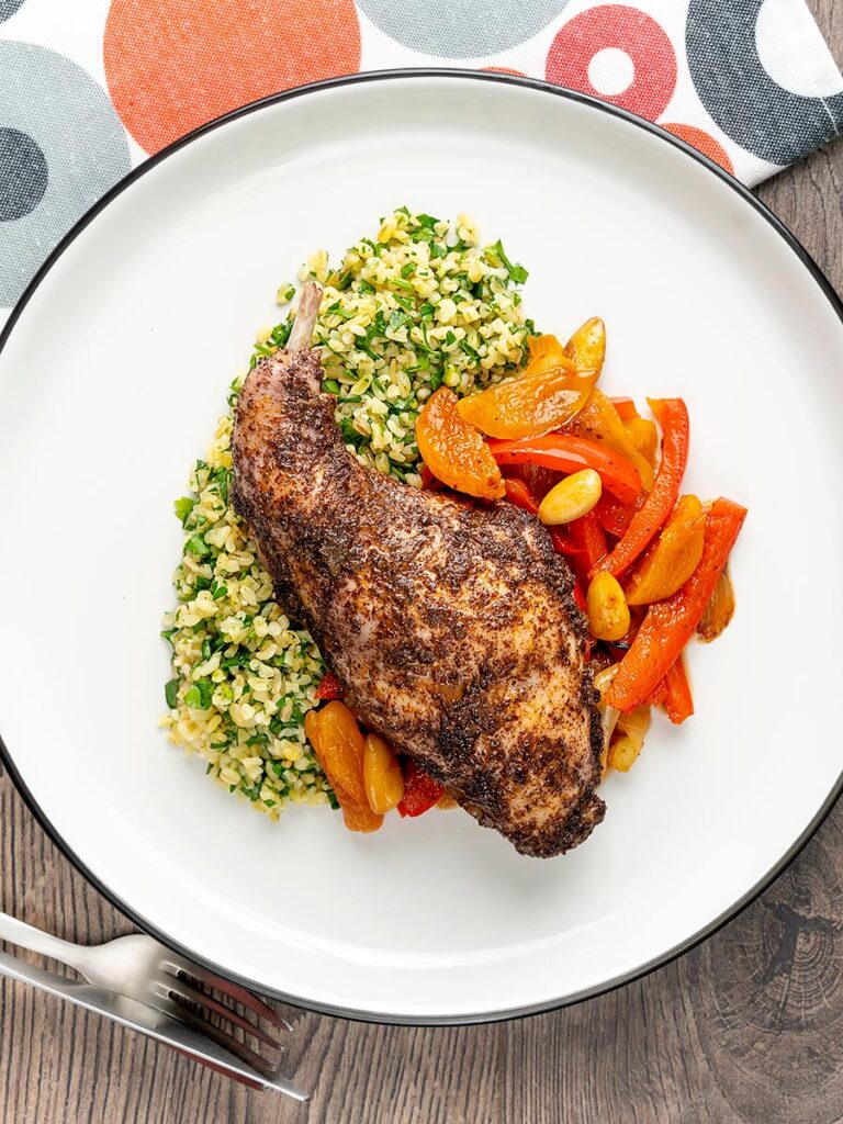 Portrait overhead image of a rabbit leg tagine with almonds and dried apricots served with herby bulgur wheat on a white plate