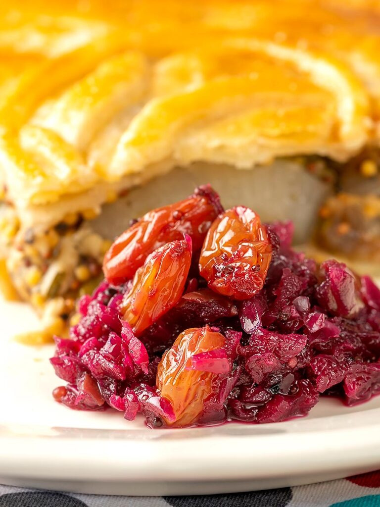 Portrait image of a red cabbage chutney with apples and raisins sat on a plate with an out of focus savoury pie in the background