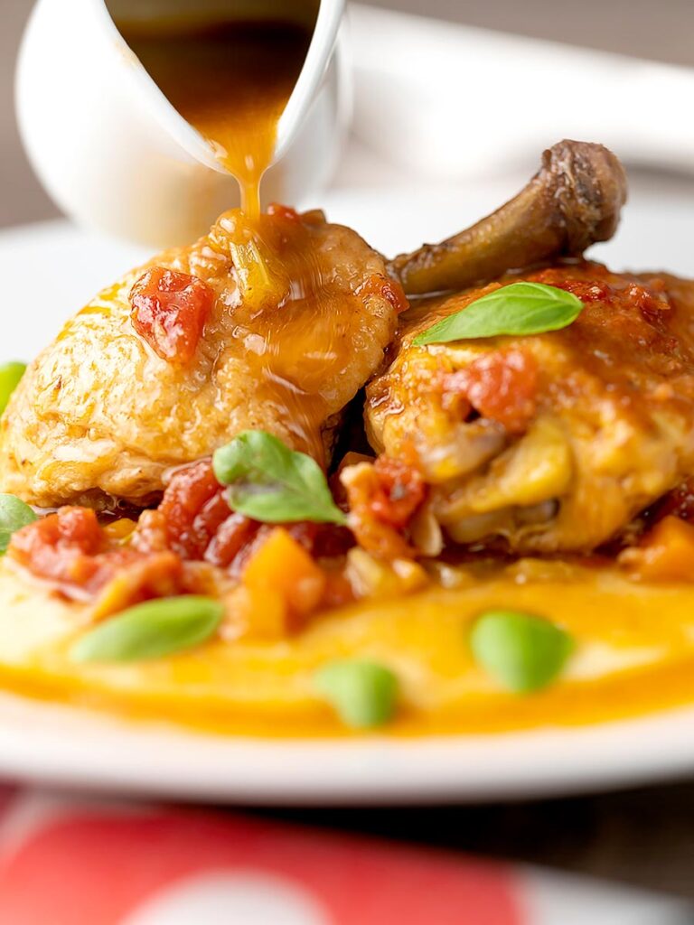 Portrait close up image of slow cooker chicken cacciatore served on a bed of polenta with extra sauce being poured from a sauce jug