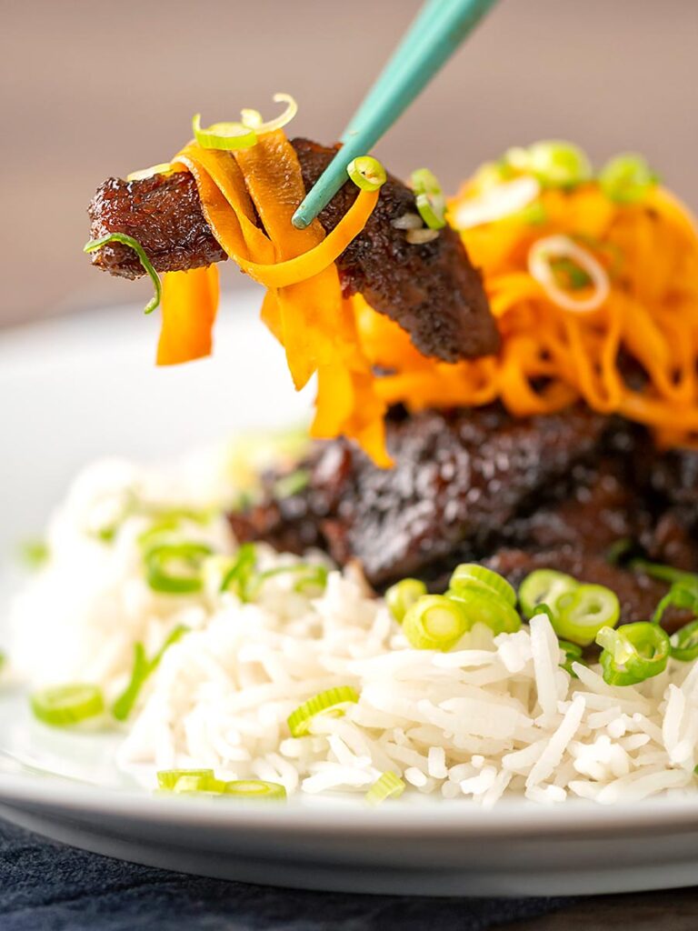 Portrait close up image of sticky crispy chilli beef with fried carrots being held in chopsticks
