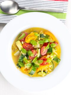 Portrait overhead image of a summer vegetable soup featuring courgettes, tomato and new potatoes served in a white shallow bowl