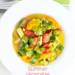 Portrait overhead image of a summer vegetable soup featuring courgettes, tomato and new potatoes served in a white shallow bowl with text overlay