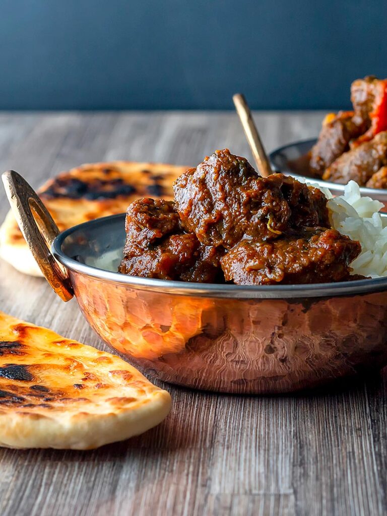 Portrait image of a beef vindaloo curry in a copper coated curry bowl served with naan bread