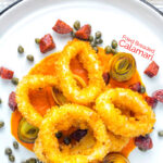 Portrait overhead image of crispy fried calamari rings that are breaded and served with romesco sauce, salami leeks, and capers with text overlay