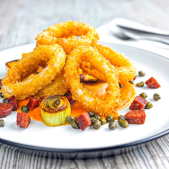 Square image of crispy fried calamari rings that are breaded and served with romesco sauce, salami leeks, and capers