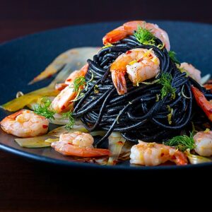 Square image of a squid ink pasta served with prawns, fennel, chilli flakes and lemon zest on a black plate