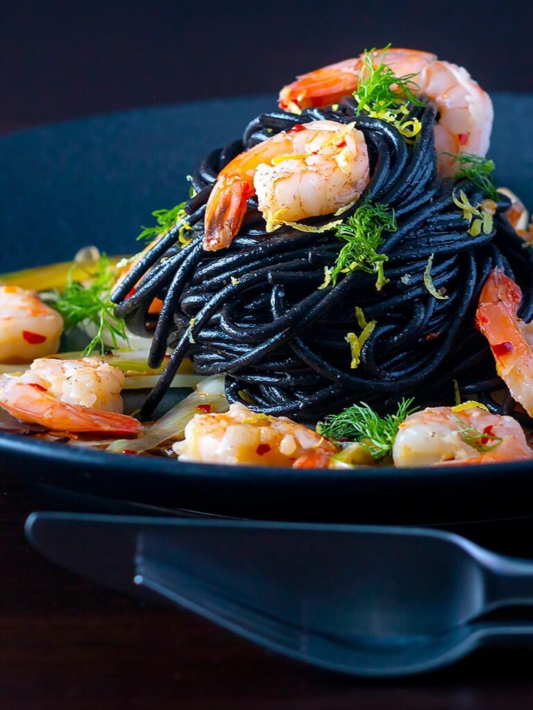 Portrait image of a squid ink pasta served with prawns, fennel, chilli flakes and lemon zest on a black plate