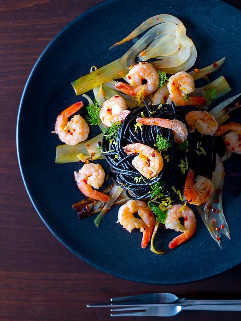 Portrait overhead image of a squid ink pasta served with prawns, fennel, chilli flakes and lemon zest on a black plate