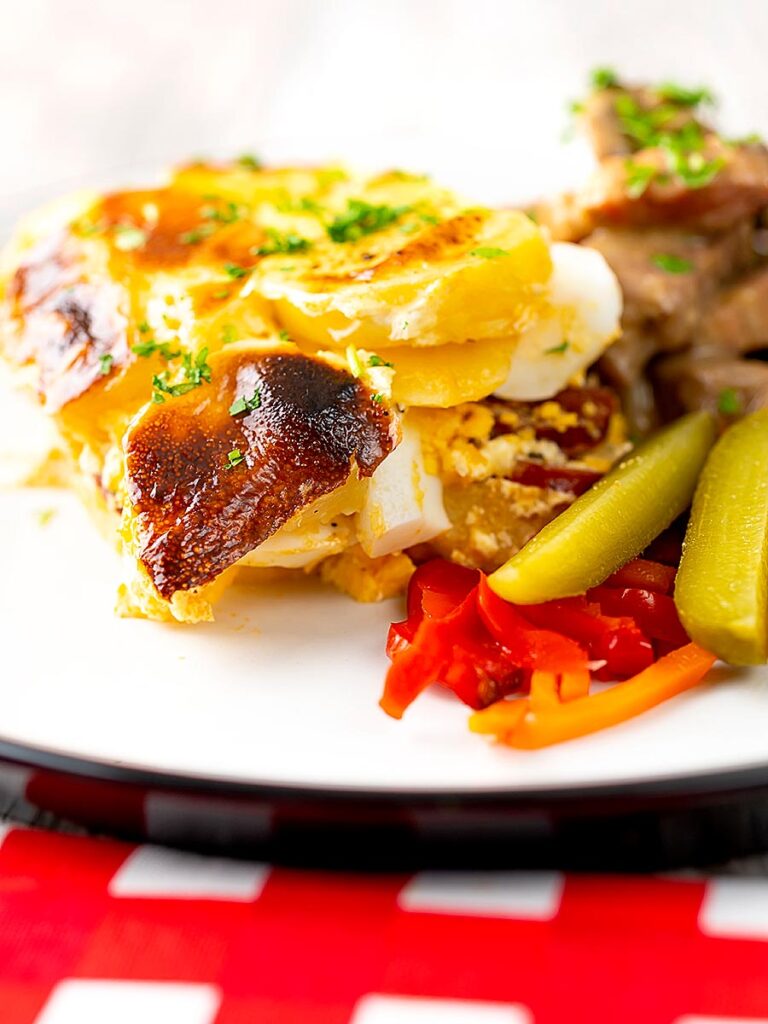 Portrait image of rakott krumpli or Hungarian layered potatoes served on a white plate with borsos tokany and pickles