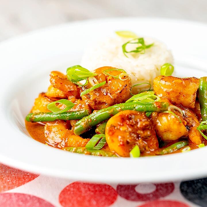 Square image of a Thai red prawn curry with green beans served in a white bowl with green onions and jasmine rice