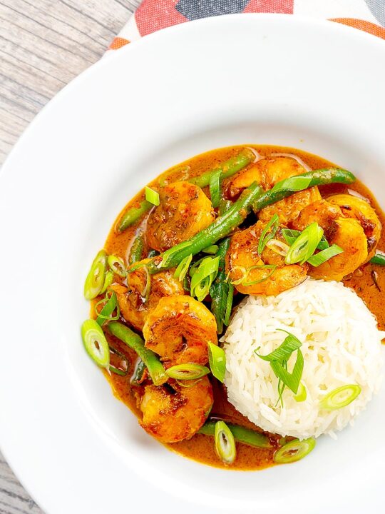 Portrait overhead image of a Thai red prawn curry with green beans served in a white bowl with green onions and jasmine rice