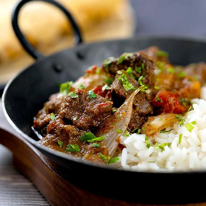 Square image of an onion rich beef dopiaza curry served in an iron karahi with rice and a coriander garnish