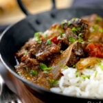 Portrait image of an onion rich beef dopiaza curry served in an iron karahi with rice and a coriander garnish and a text overlay