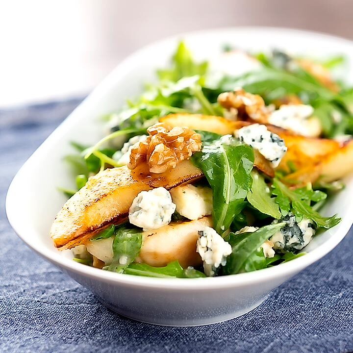 Square image of a pear and blue cheese salad with rocket (arugula) and walnuts served in a white bowl