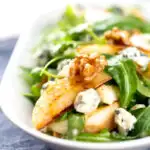 Portrait image of a pear and blue cheese salad with rocket (arugula) and walnuts served in a white bowl with a text overlay