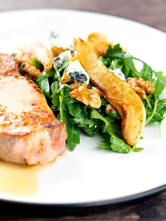 Portrait image of a pear and blue cheese salad with rocket (arugula) and walnuts served as a side dish to a pork chop