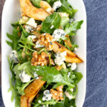 Portrait overhead image of a pear and blue cheese salad with rocket (arugula) and walnuts served in a white bowl with a text overlay