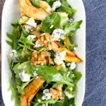 Portrait overhead image of a pear and blue cheese salad with rocket (arugula) and walnuts served in a white bowl with a text overlay