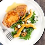 Portrait overhead image of a pork loin steak in a cider sauce served on a white plate with a pear and blue cheese salad