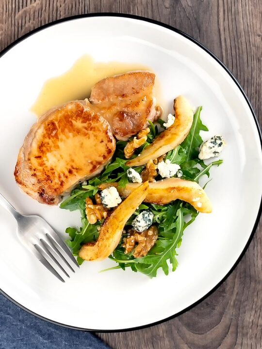 Portrait overhead image of a pork loin steak in a cider sauce served on a white plate with a pear and blue cheese salad