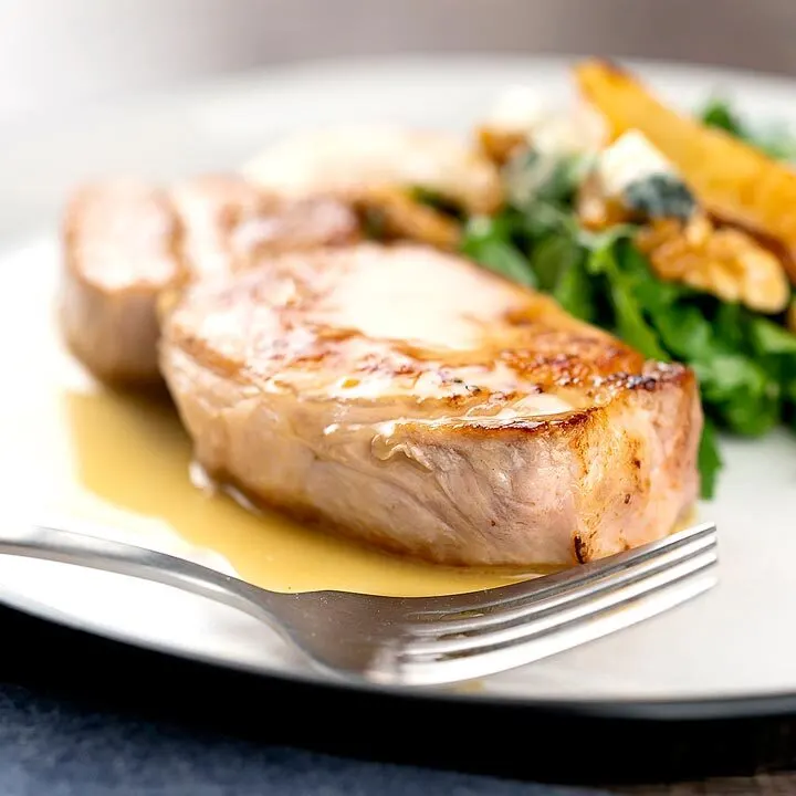 Square image of a pork loin steak served on a white plate with a cider sauce and side salad