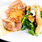 Portrait close up image of a pork loin steak in a cider sauce served on a white plate with a pear and blue cheese salad