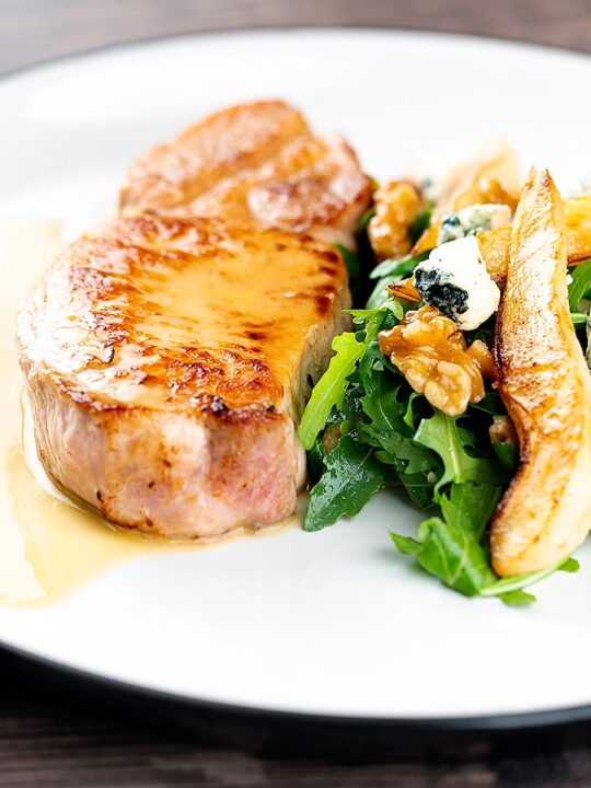 Portrait close up image of a pork loin steak in a cider sauce served on a white plate with a pear and blue cheese salad