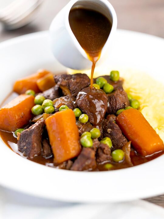 Portrait image of extra gravy being poured over a slow cooker venison stew or casserole served with peas, carrots and mashed potato