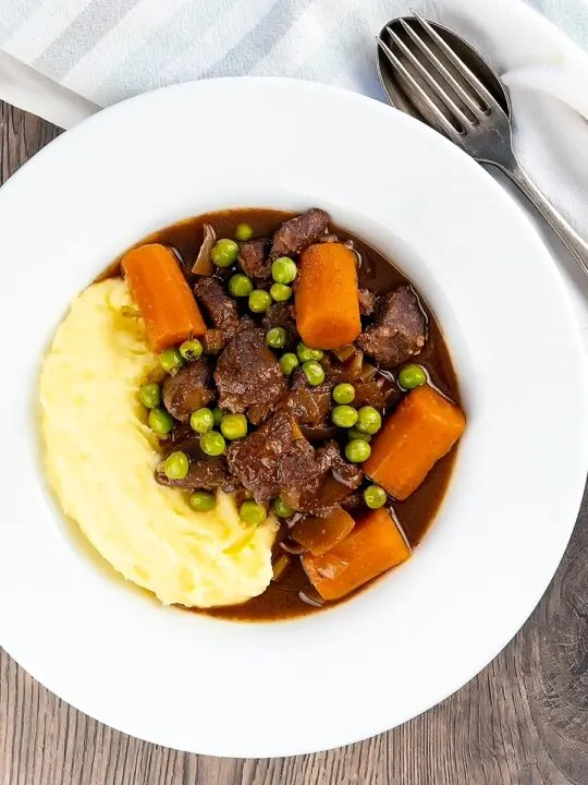 Portrait over head image of a slow cooker venison stew or casserole served with peas, carrots and mashed potato
