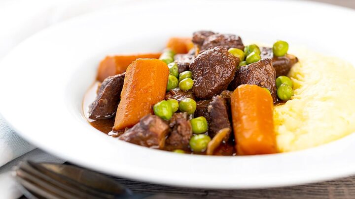 Landscape image of a slow cooker venison stew or casserole served with peas, carrots and mashed potato