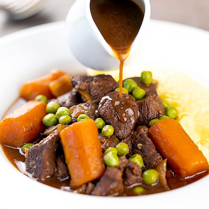 Square image of extra gravy being poured over a slow cooker venison stew or casserole served with peas, carrots and mashed potato