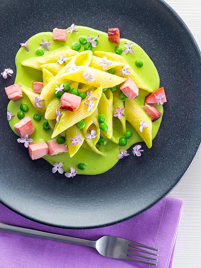 Portrait overhead image of ham and pea pasta using penne rigatoni and edible lilac flowers as garnish