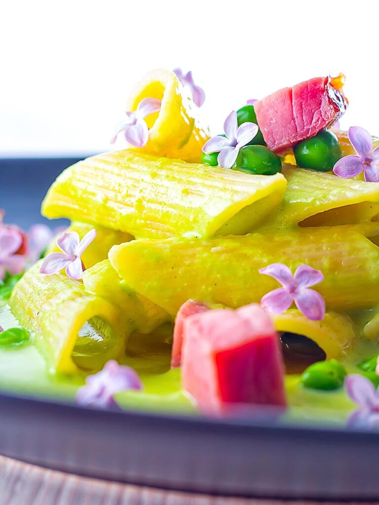 Portrait close up image of ham and pea pasta using penne rigatoni and edible lilac flowers as garnish