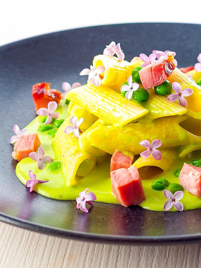 Portrait image of ham and pea pasta using penne rigatoni and edible lilac flowers as garnish