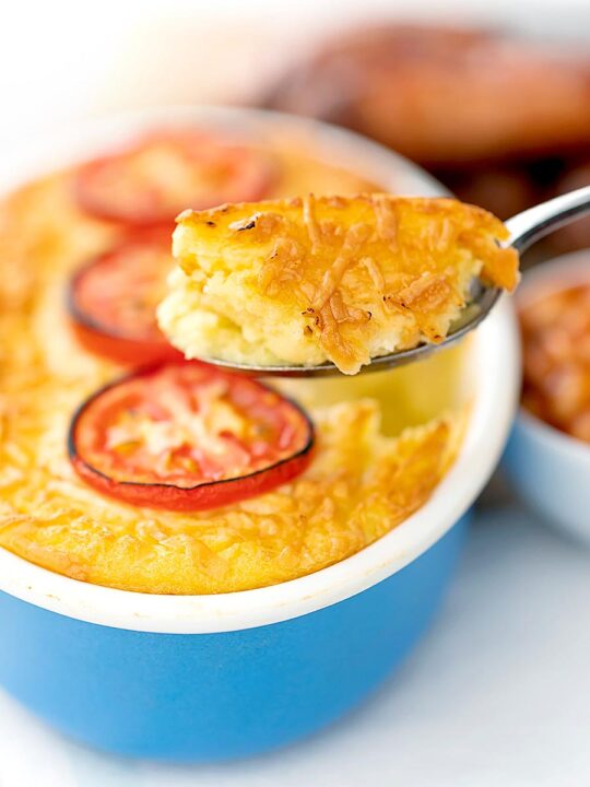 Portrait image of cheese and potato pie bake topped with tomato slices with a spoon taking out a piece