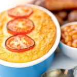 Portrait image of cheese and potato pie bake topped with tomato slices and served with sausages and baked bean
