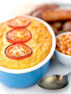 Portrait image of cheese and potato pie bake topped with tomato slices and served with sausages and baked bean