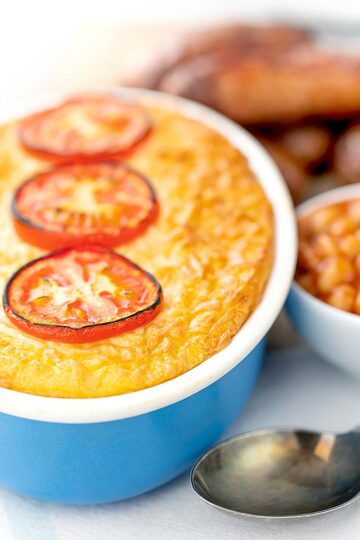 Portrait image of cheese and potato pie bake topped with tomato slices and served with sausages and baked bean