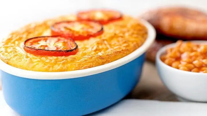 Landscape image of cheese and potato pie bake topped with tomato slices served with baked beans and sausages