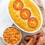 Portrait overhead image of cheese and potato pie bake topped with tomato slices and served with sausages and baked beans