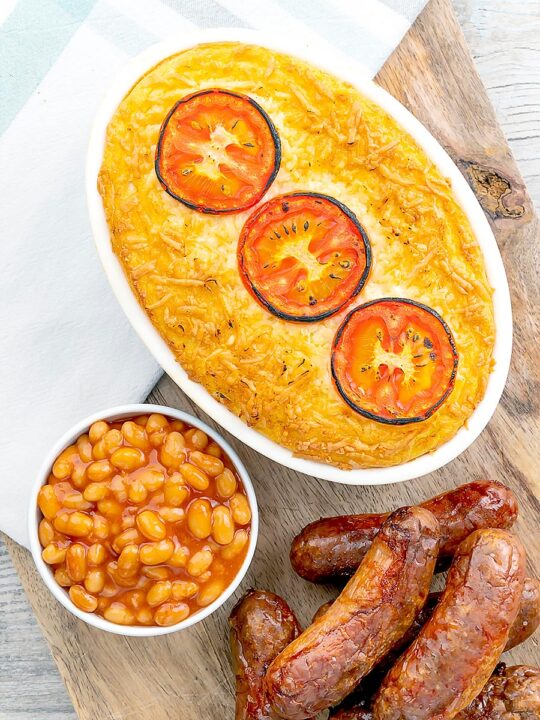 Portrait overhead image of cheese and potato pie bake topped with tomato slices and served with sausages and baked beans