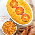 Portrait overhead image of cheese and potato pie bake topped with tomato slices and served with sausages and baked beans featuring a text overlay